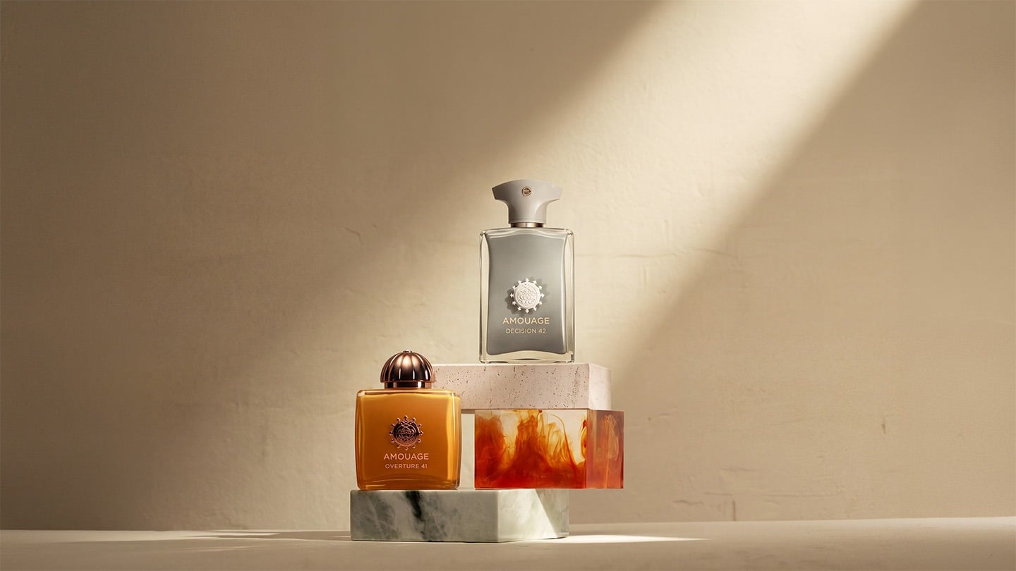Two Amouage fragrance bottles, Decision 42 and Overture 41, displayed on sculptural stone and amber-toned blocks against a warm beige background with soft sunlight